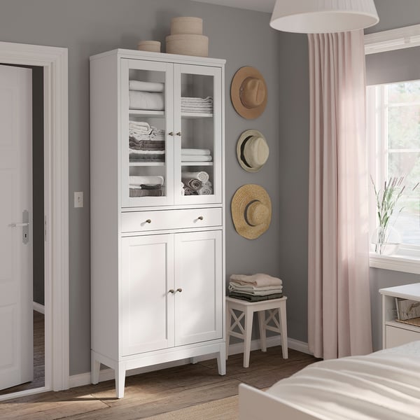 White cabinet with glass doors, drawers, and hats display. Organised storage in bedroom.