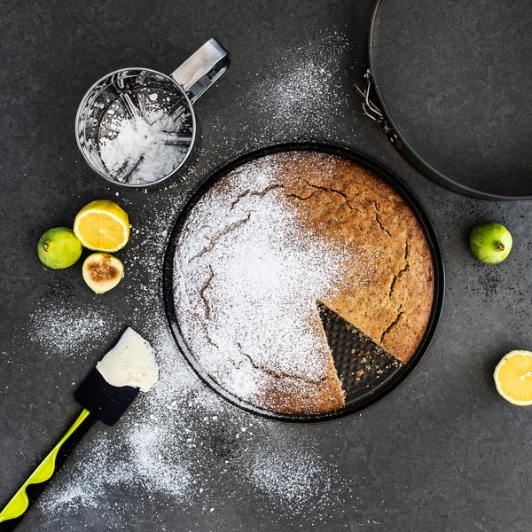 A round black cake on a tray dusted with icing sugar, surrounded by lemons, limes, a sifter, and a spatula.