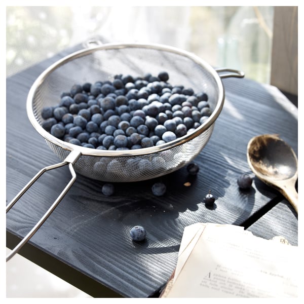 A stainless steel strainer filled with blueberries sits on a dark wooden surface. A spoon and an open book lie nearby.