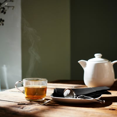 Glass mug with golden tea, steam rising, IDEALISK tea infuser on napkin next to white teapot on wooden table.