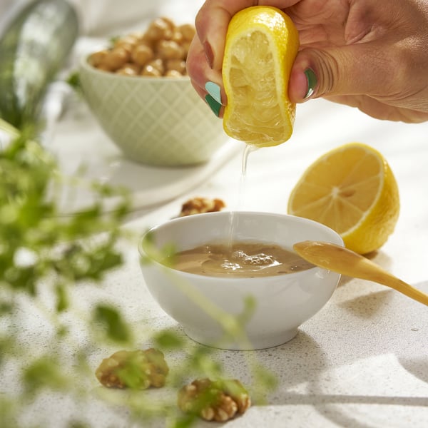 Person squeezes lemon into a white bowl on a table with walnuts and greens.