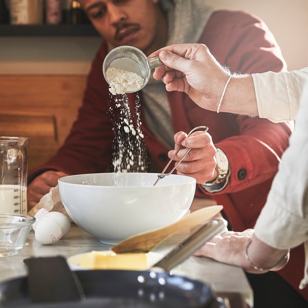 Person pours flour into mixing bowl using measuring cup. Bowl, ingredients, and kitchen tools are on the table.