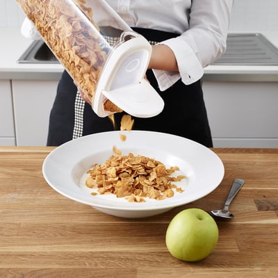 Person pours cereal from a transparent IKEA container into a bowl on a wooden counter. An apple and spoon are nearby.