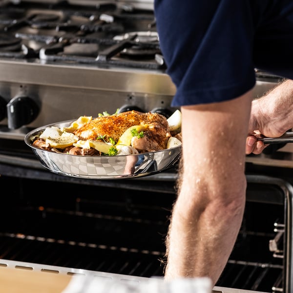 Person holding a glass frying pan with cooked chicken and vegetables, placing it into an oven.
