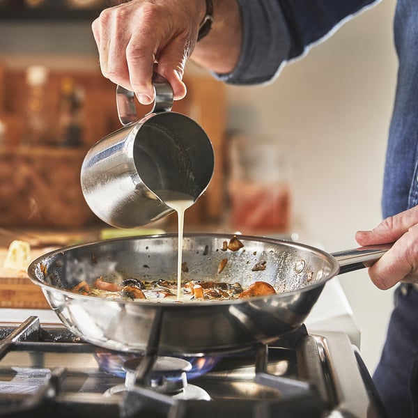 Person cooking on stove, pouring liquid from metal pitcher into frying pan with food.