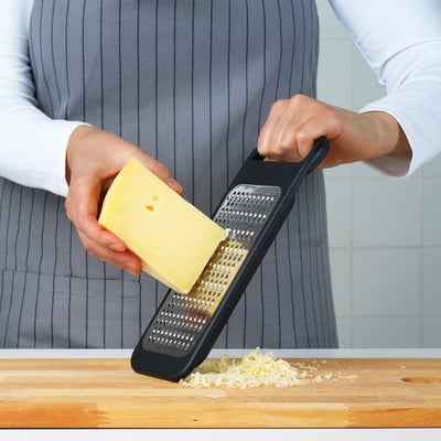 Person grates cheese on black kitchen tool with steel surface and stable base.