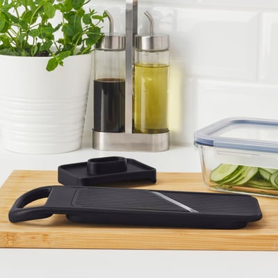 Black mandoline slicer on bamboo board, near oil bottles, herbs, and food container on white countertop.
