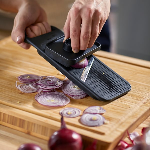 Person slicing onions on a wooden cutting board using an IKEA 365+ mandoline slicer, black. The tool is positioned to slice the onions uniformly.