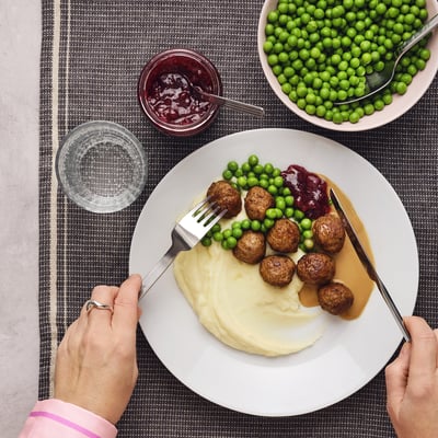 Person holding plate with meatballs, mashed potatoes, peas, and jam. Bowls and cups surround the plate.