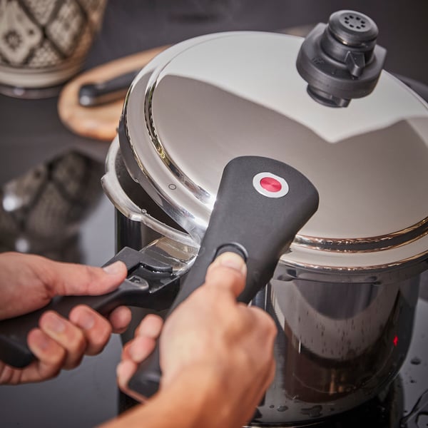 A pair of hands operates a silver IKEA 365+ pressure cooker, gripping black handles, ensuring safety and efficiency in cooking.