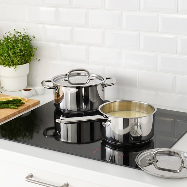 Modern kitchen with saucepans on induction hob, one with lid, other with cream. Cutting board with veggies and herb, white tiled wall.