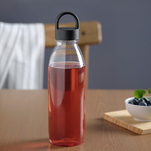Clear bottle with dark red liquid and black lid handle on wooden table near a white bowl with greenery.