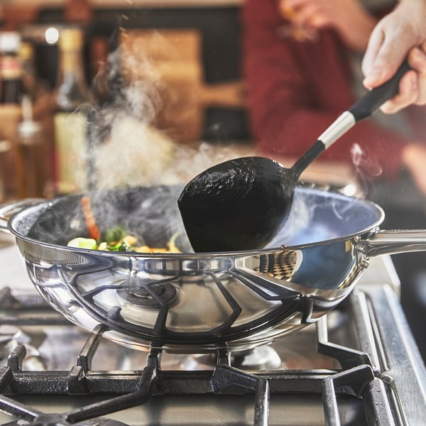 Cooking in progress: a hand stirs vegetables in a wok on a gas stove. Steam rises from the pan, indicating active cooking.