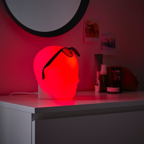ISKÄRNA lamp glows red on desk, surrounded by personal items, with circular mirror, showing creative design with sunglasses shape.