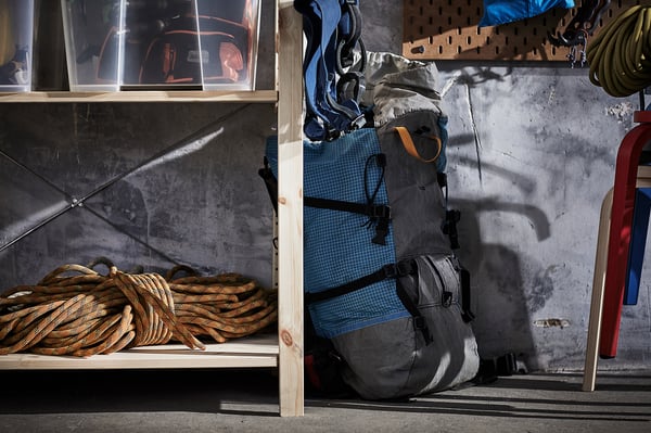 A storage area with IVAR shelves, holding a coiled rope, a grey and blue backpack, and various equipment, against a concrete wall.