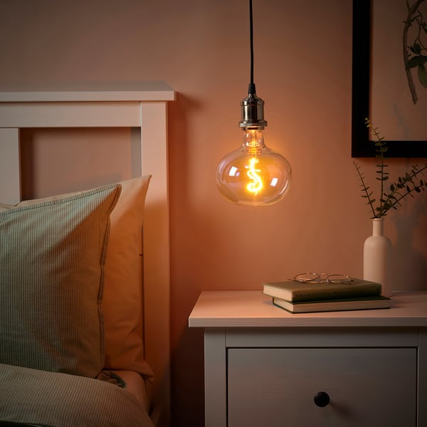 JÄLLBY pendant lamp with warm led over bedside table, multicoloured bulb, book, glasses, vase with greenery. Peach wall.
