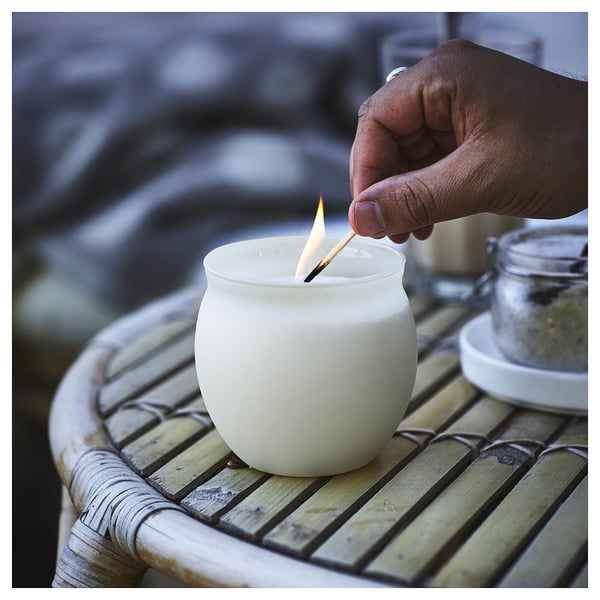 Hand lights round white JÄMLIK scented candle on bamboo table. Candle sits on glass base, emits warm light.