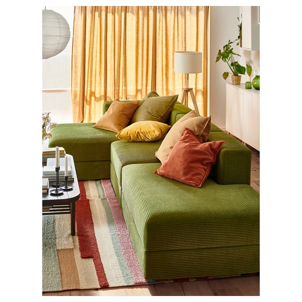 Bright living room with green JÄTTEBO modular sofa, colourful pillows, wooden floor, and large window with yellow curtains.