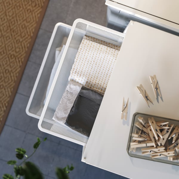JONAXEL white storage basket on floor, textured pattern, with cloth and clothespins nearby.