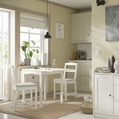 Bright kitchen with white dining set, green wall, large window, plant, pendant light, and beige rug.