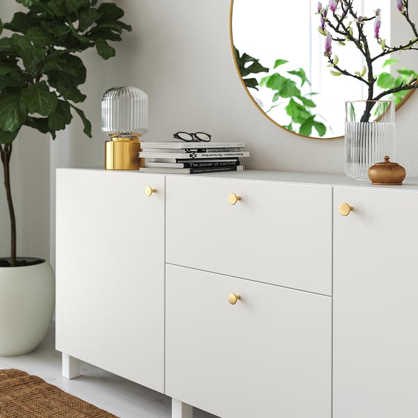 White cabinet with gold knobs, holding books and decorative items. A large plant is nearby.