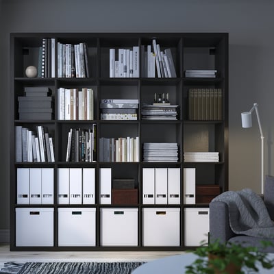 KALLAX black bookshelf, organises books and storage boxes. Tall, fits against dark wall, with chair.