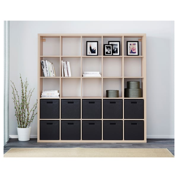 A wooden KALLAX shelving unit with black storage boxes and open shelves displaying books and framed photos.
