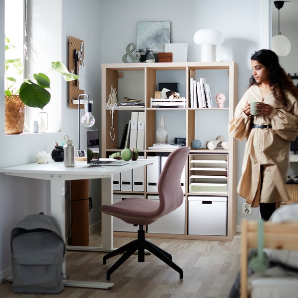 Home office with KALLAX shelf, person with mug, plant by window.