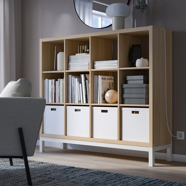 KALLAX light wood bookshelf with grey chair, filled with white and grey storage boxes and books, emphasising its size.