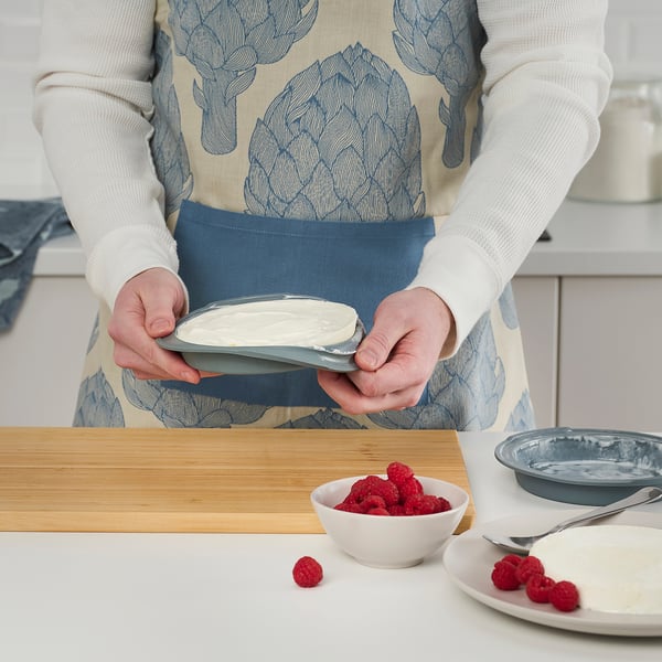 A person is holding a baking mould, preparing to make a layered cake or frozen dessert. Raspberries and icing are visible nearby.