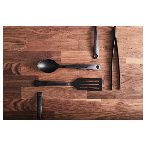 Four black kitchen utensils on wooden surface. Includes ladle, spatula, spoon, and tong. Dark wood background with visible grain patterns.
