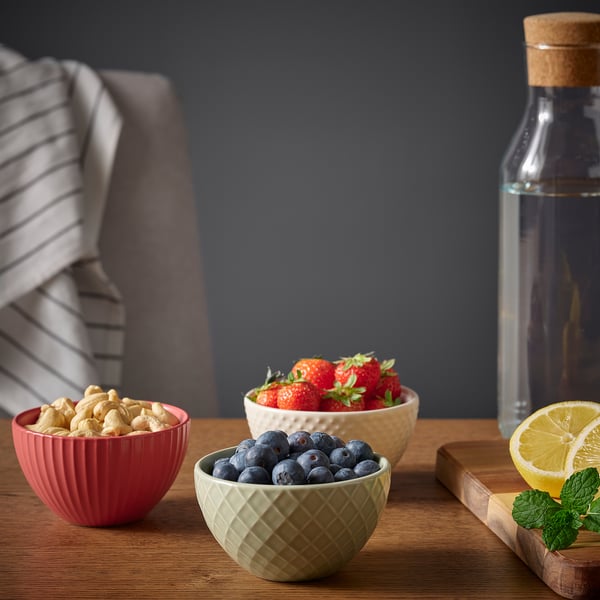 Three colourful bowls filled with cashews, strawberries, and blueberries on a wooden table with a striped cloth, a glass bottle, and a lemon slice.