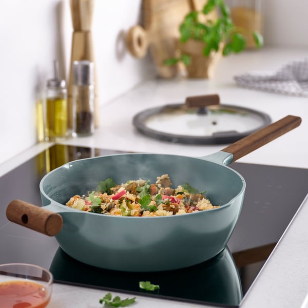 Grey-turquoise sauté pan on stovetop with wooden handles, glass lid, colourful food.