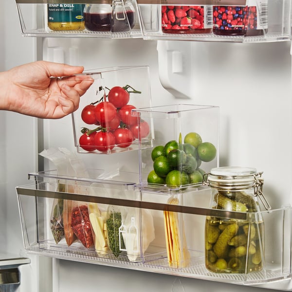 Hand reaches for cherry tomatoes in clear fridge organiser with limes, pickles, and cheeses.