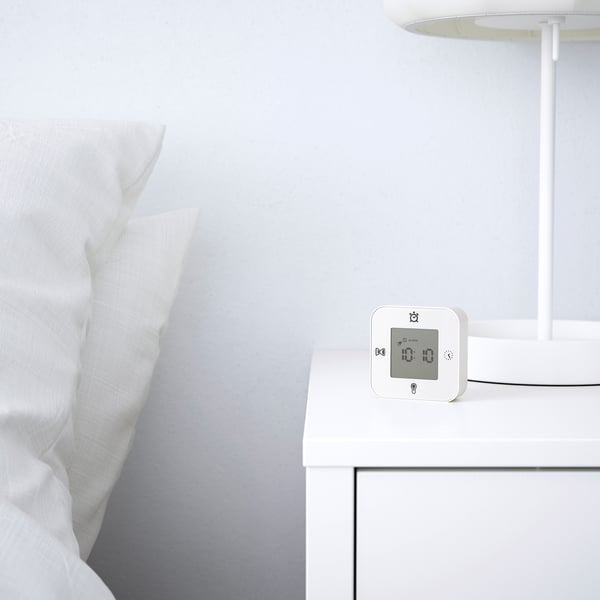 A white alarm clock sits on a bedside table, displaying time and temperature.