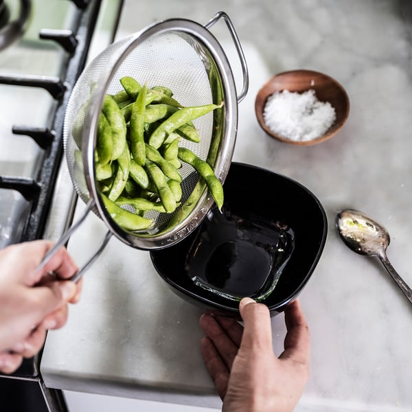 A person pours bright green snap peas from a silver mesh strainer into a black bowl. A small bowl of salt and a spoon are on the counter.