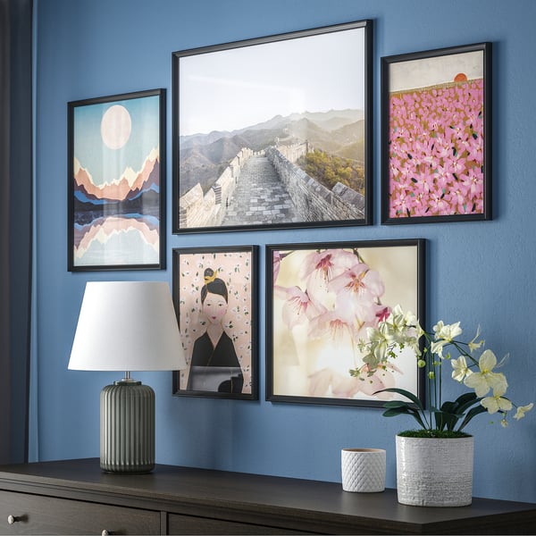 Display of five black-framed colorful pictures on blue wall, lamp, and plant on table.