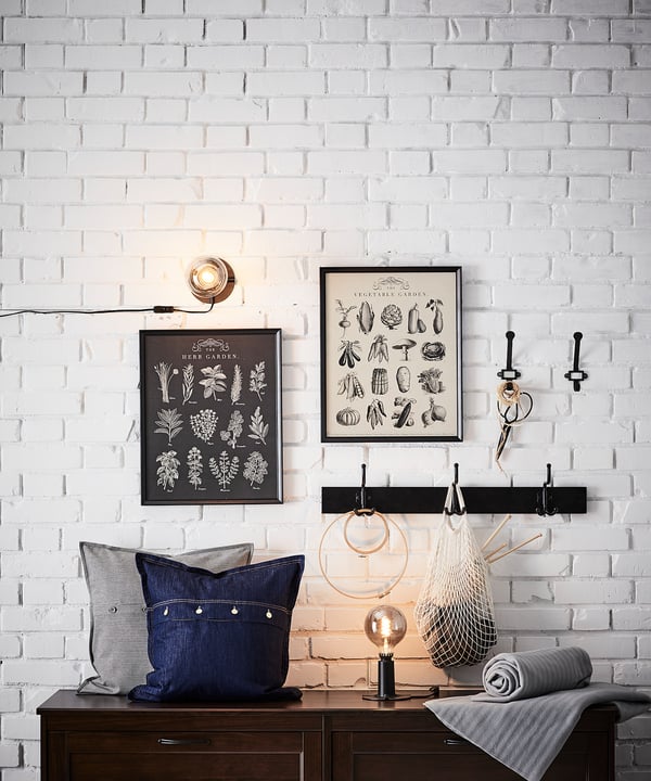 Wall-mounted light and posters on a white brick wall. Below, cushions on a dark furniture piece.