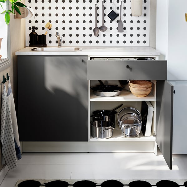 Modern kitchen with open drawer showing pots and pans. Dark grey cabinet, marble worktop, and polka dot wall. Sink and storage nearby.