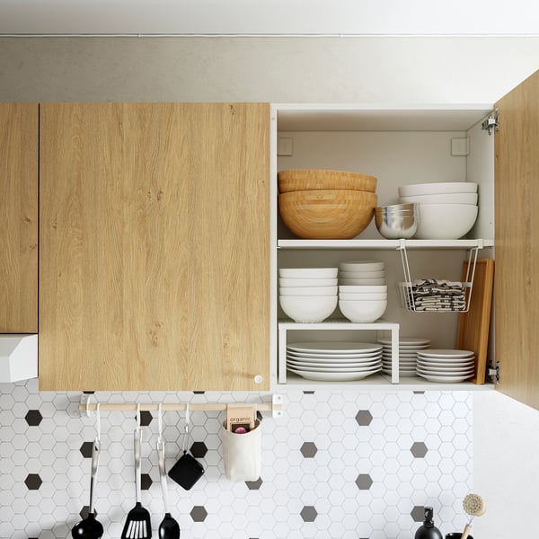 Open kitchen cabinet showing organised dishes: bowls, plates, and cutlery. Sleek wooden finish, white tiled backsplash.