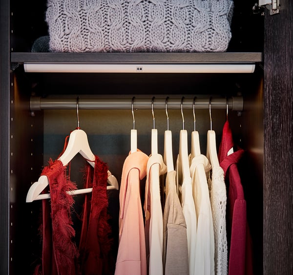 Closet with KOMPLEMENT clothes rail hanging various coloured clothes. Red, beige, and white garments on wooden hangers. Light above.