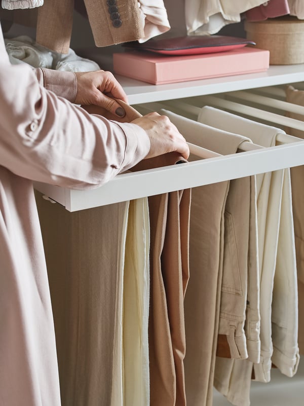 A person organises beige pants using KOMPLEMENT trouser hanger, white, steel, and fiberboard, in a wardrobe.