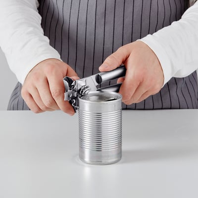 A person in a striped shirt uses a can opener to open a metal can on a white surface.