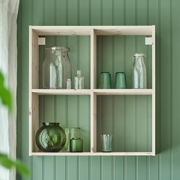 Wall-mounted shelf with clear and green glass jars in compartments against green wall.