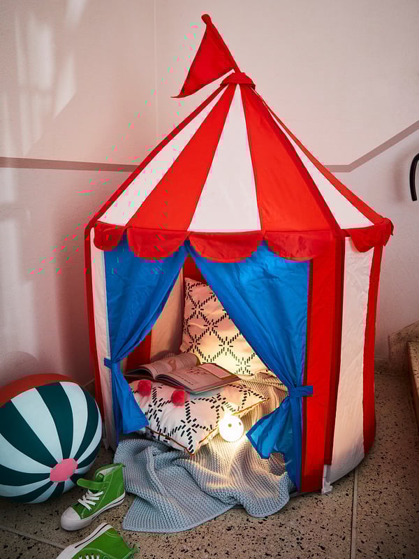 Bright kids play tent shaped like a circus tent, red and white striped top with blue walls, cosy inside with pillows and a small lamp.