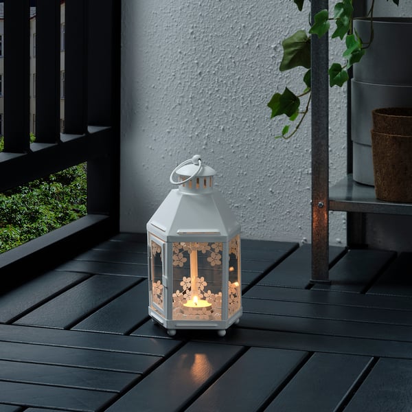 A small white lantern sits on a dark wooden table, casting warm light through its floral pattern.