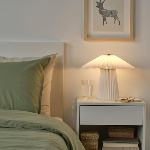 A cozy bedroom with a KUDDLAVA lamp on a small table, featuring a green pillow, books, and a deer picture.