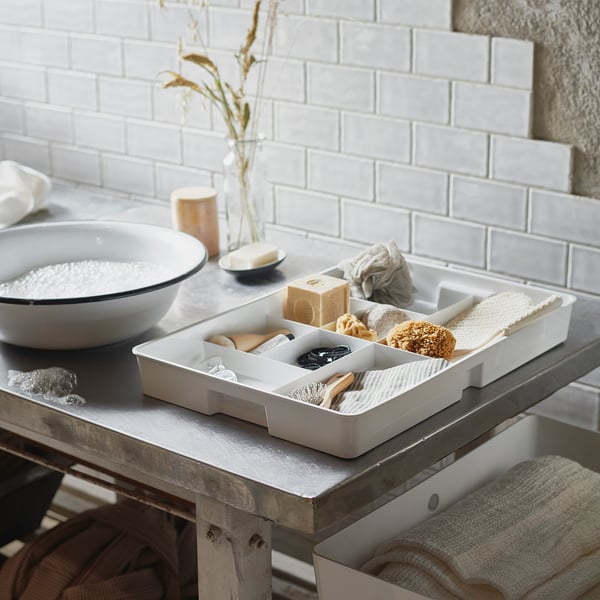 White KUGGIS organiser tray on a countertop, contains small bathroom items, against a tiled wall.