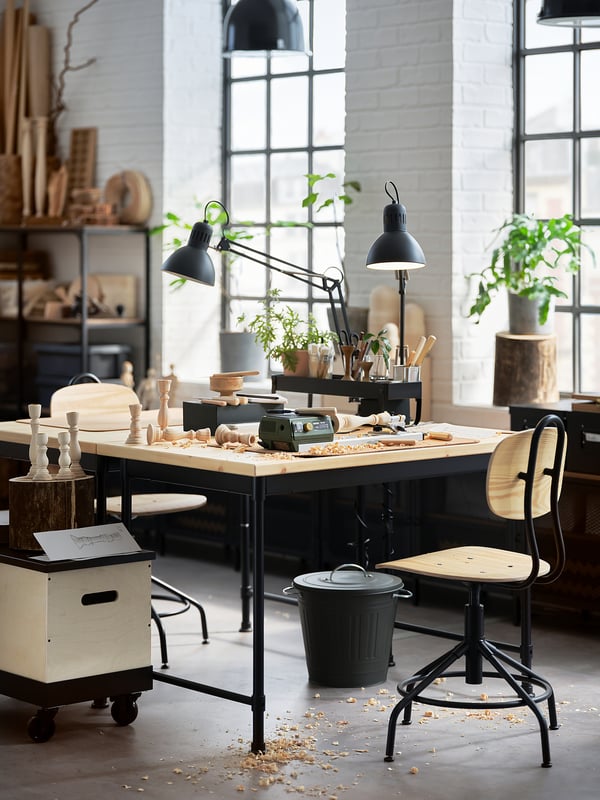 A workspace featuring KULLABERG black desks with wooden tops, housing tools and plants, with large windows and a minimalist design.