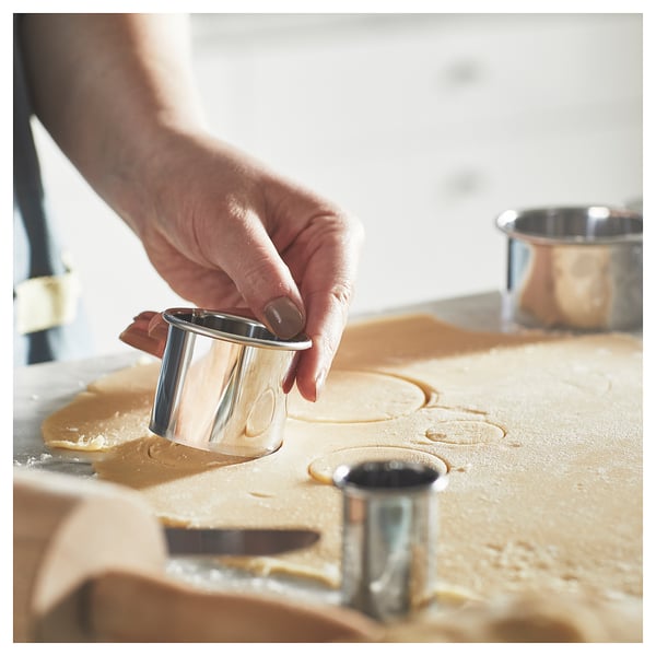 A person uses a silver cookie cutter to make shapes in dough. Several cutters lie on the floured surface, ready for use.